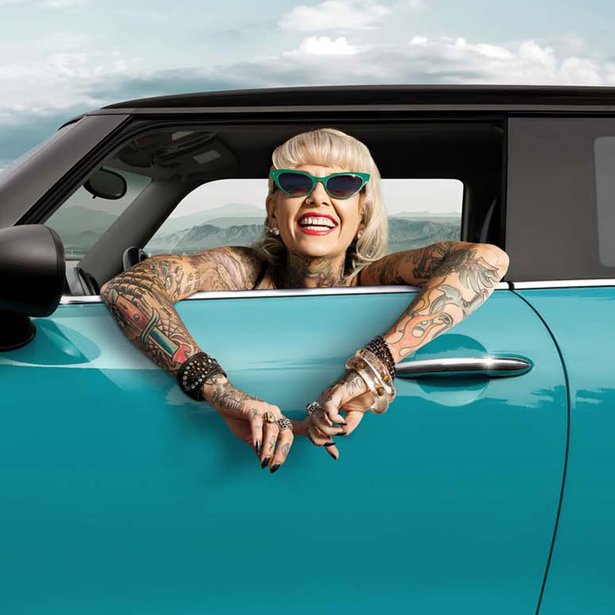 Eine tätowierte Frau mit grüner Sonnenbrille schaut lachend aus einem BMW Mini, die Arme liegen lässig über der Fahrertür.